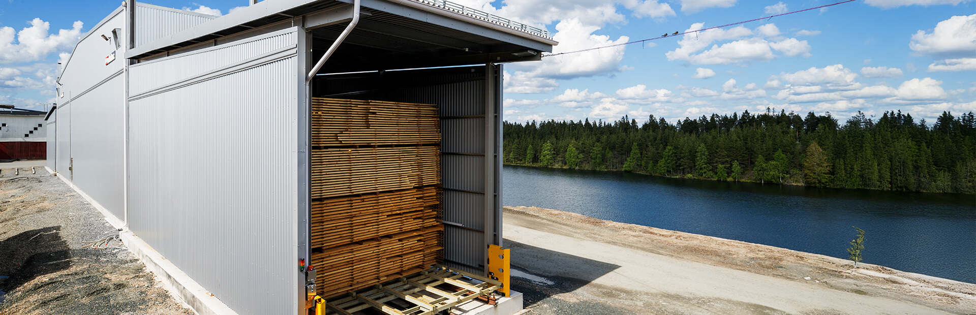 Silverfärgad Valutec kanaltork FB med virkespaket lastade i öppningen. I bakgrunden syns sjö och skog under blå himmel med moln.