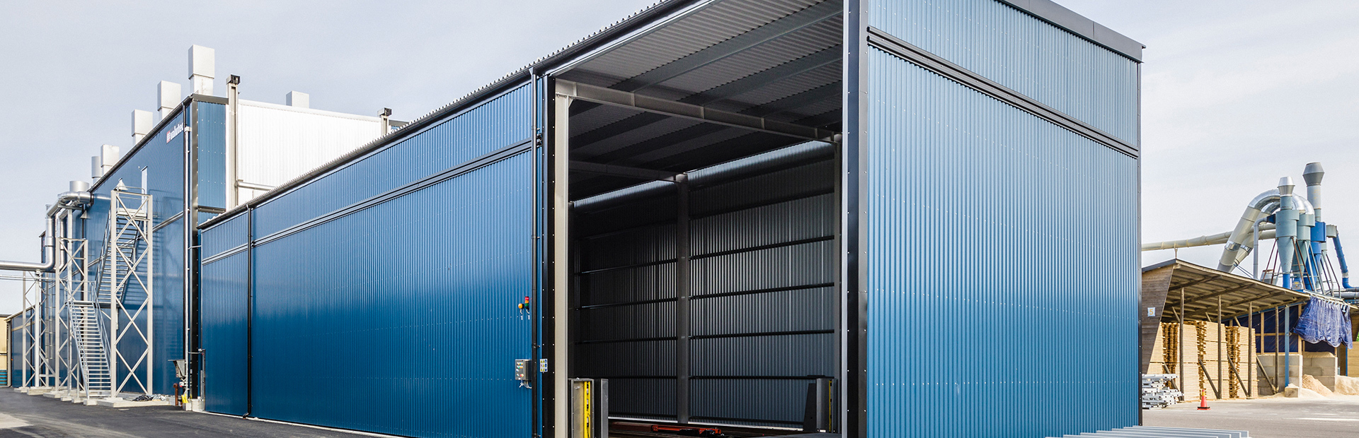 Blå Valutec kanaltork TC med öppen port, sedd från sidan. Stålkonstruktion och kringliggande sågverksmiljö i bakgrunden.