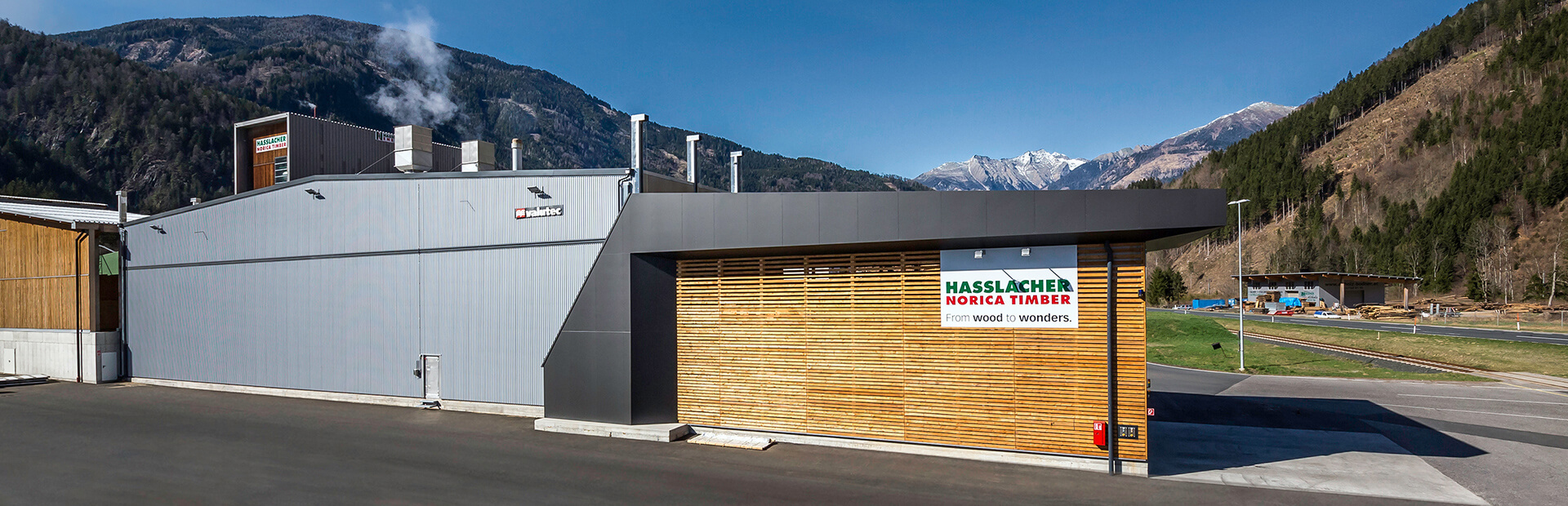 Valutec OTC-kanaltork vid Hasslacher sågverk, byggnad i grå plåt med träfasad och berg i bakgrunden.