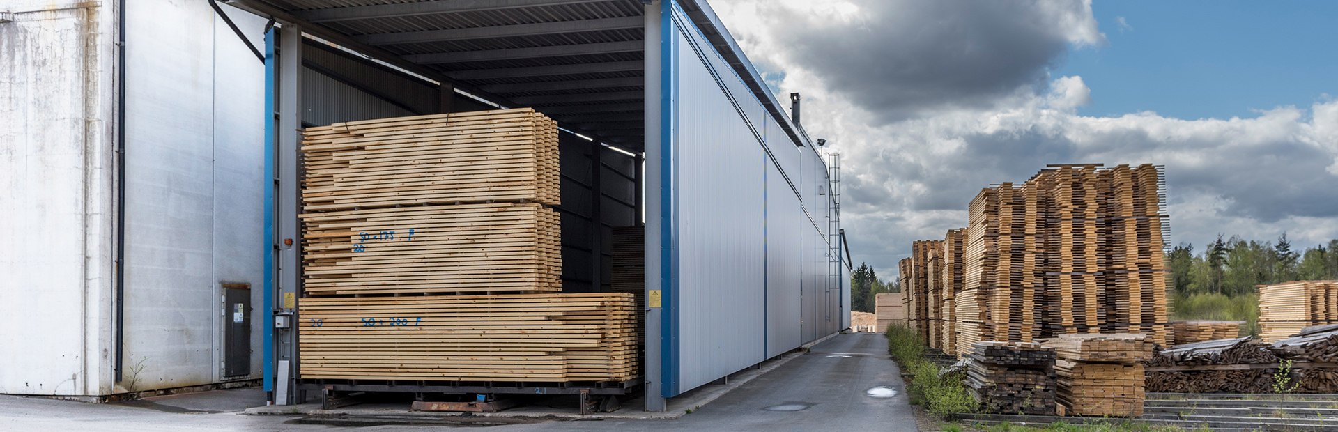 Kanaltork OTC från Valutec med blå fasad, lastad med virkespaket. Staplade virkeslager syns utomhus i bakgrunden.